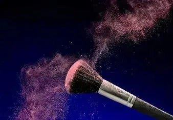 Makeup brush dispersing pink powder against dark blue background